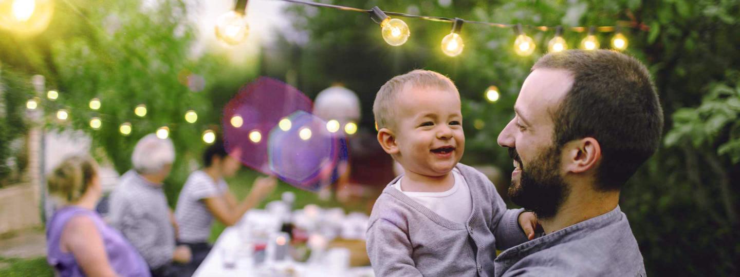 Vater mit kleinem Kind auf dem Arm freut sich bei einer Familienfeier im Garten über den günstigen Strompreis